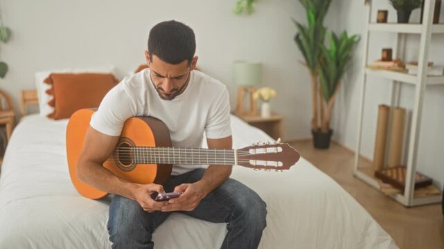 Man, seated on bed plays acoustic guitar while looking at smartphone in studio; quiet contemplation.