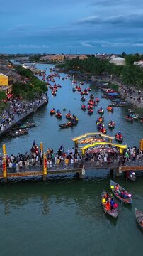 Vertical aerial drone video of Hoi An Ancient Town at dusk, Vietnam, featuring colorful lantern boats, crowded riverside promenade, and vibrant tourism atmosphere along the Thu Bon River.
