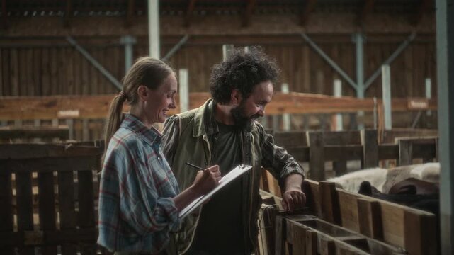 Female manager taking notes on clipboard and discussing animal health with bearded male farmer as he checking on cows inside cattle pen in barn