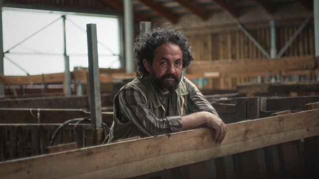 Middle-aged bearded male farmer in vest and plaid shirt leaning on wooden cattle pen fence and looking at camera inside rustic agricultural barn. Video portrait