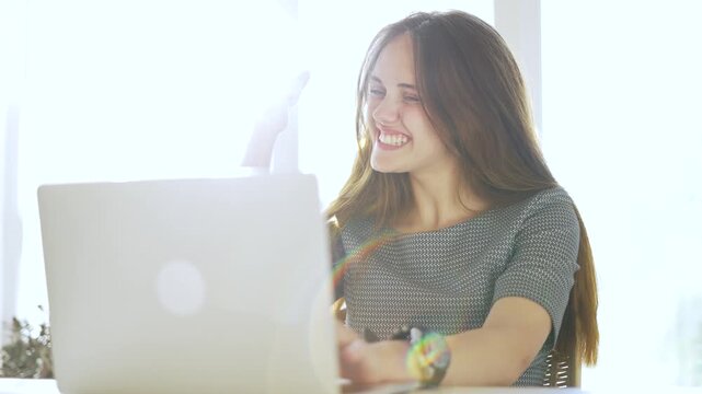 Young woman smiles while using laptop by bright sunny window. Smiling girl works on computer at home office. Happy female enjoys remote work. Sunlight fills room as she types and laughs softly.
