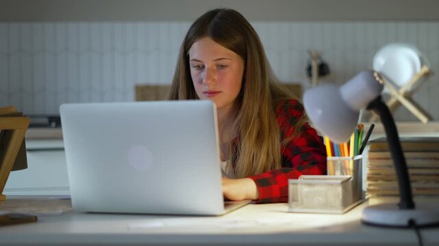 Young teenager sits at home desk typing on silver laptop. Female student is studying online now. Cheerful girl uses computer for internet education. Teenager enjoys studying via laptop at home.