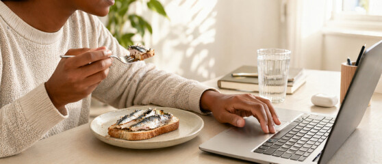 Person eating sardine toast while working on laptop at home office © dvoevnore