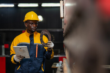 Fototapeta premium Worker wears yellow hard hat and blue work uniform. Holds metal ring and tablet computer. Gloves cover hands while inspecting tools. Industrial workspace features machinery and overhead lights