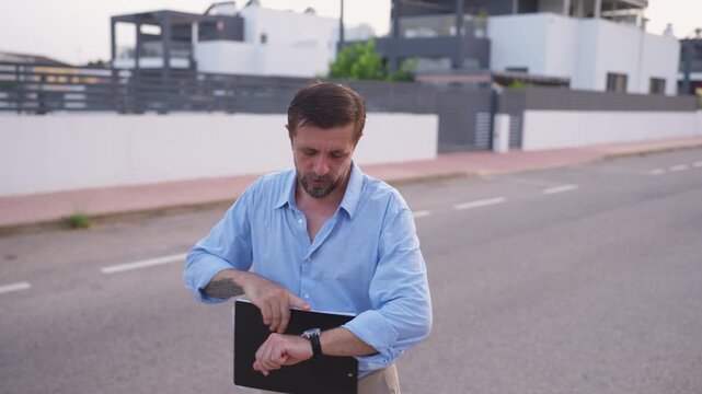Anxious male wait outdoors, impatiently check watch while fidgeting with clipboard and documents, expressing frustration, stress, impatience, delay, and concern about a missed appointment or deadline