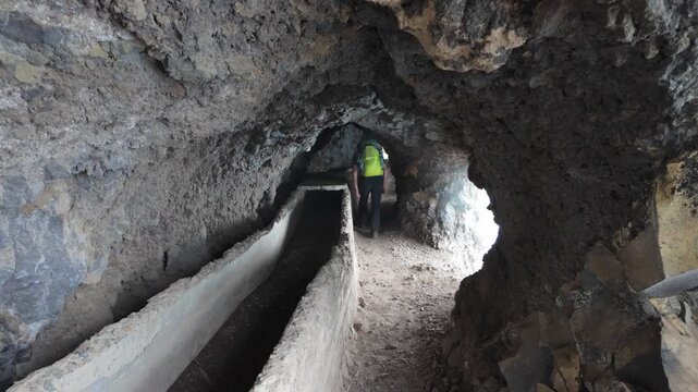 A lone hiker navigates the rocky water tunnels of "Las Ventanas de G&uuml;&iacute;mar" on Tenerife, revealing the rugged interior with water canals and windows at the side. Emphasizes exploration and adventure.