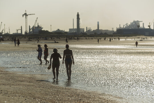 Silhouettes of two people walk on the beach at Dunkirk at low tide, the wet sand reflecting the light while the distant port appears on the horizon.