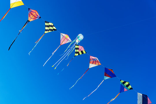 A vibrant string of colorful kites including diamond and a jellyfish shape flies against a clear blue sky.