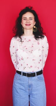 Attractive playful Caucasian woman holding hands behind back winking eye with flirty smile touching chest on bright red studio background, cheerful mood flirtatious gesture concept stock video clip