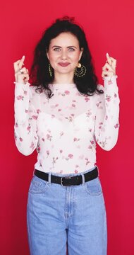 Attractive Caucasian woman crossing fingers for good luck looking at camera with hopeful joyful smile on bright red studio background, wishing success superstitious gesture concept stock video clip