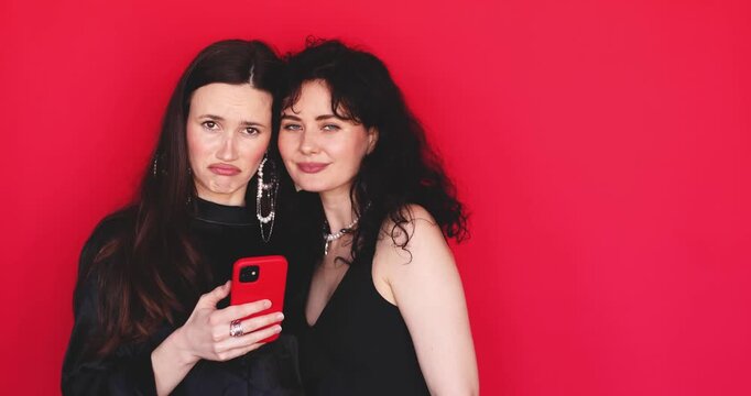 Two attractive Caucasian women looking at smartphone screen with sad expression feeling sorry and disappointed face on bright red background, bad news reaction online content stock video clip