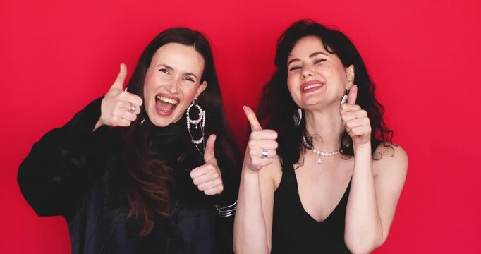 Two attractive Caucasian women showing double thumbs up gesture with cheerful smiling faces looking at camera on bright red studio background, enthusiastic approval concept stock video clip