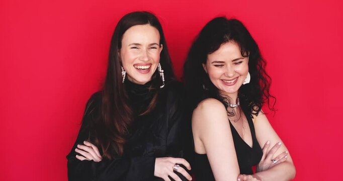 Two attractive Caucasian women standing with arms crossed starting to laugh out loud with genuine joyful emotion looking at camera on bright red background, cheerful friendship concept stock video 