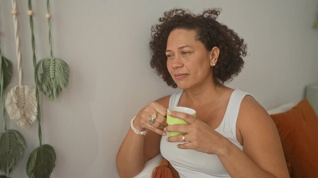 Woman holding a green mug and smiling while seated on a bed in a cozy bedroom with pillows and macrame wall art; calm contentment.