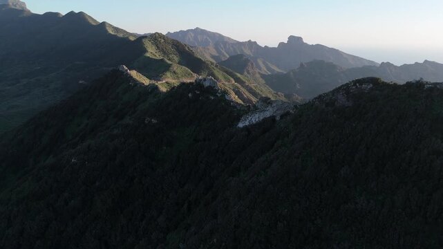 Aerial pan left drone shot revealing the dramatic, rugged landscape of Cumbre del Carrizal on Tenerife, Canary Islands, at sunset. Winding road on a mountain ridge and vast ocean in the distance.