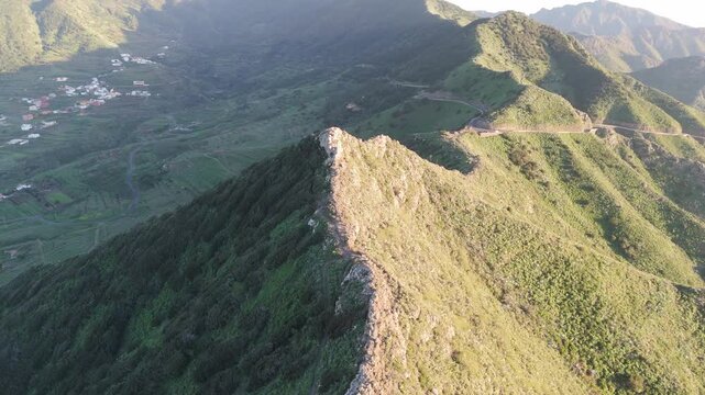Aerial drone push in shot revealing the rugged landscape of Cumbre del Carrizal mountain on Tenerife, Canary Islands, bathed in the warm light of sunset. Lush green valleys and a village are visible.