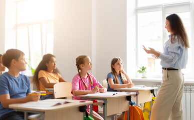 Obraz premium Female teacher explaining lesson to attentive children seated at individual desks in bright multicultural classroom, encouraging participation and active learning