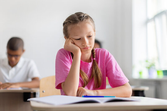 Tired schoolgirl resting her head on one hand beside an open notebook during class, expressing boredom, fatigue, or difficulty concentrating in a bright school setting