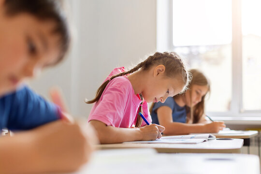 Focused schoolgirl with braided hair writing in open notebook at her desk during a quiet classroom lesson, while classmates complete assignments in the background
