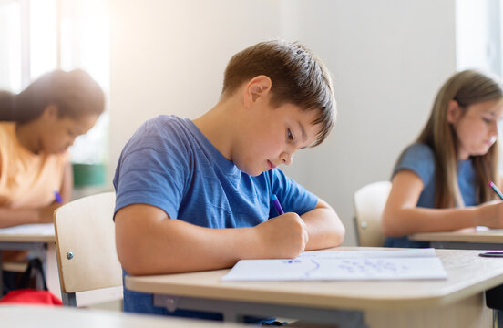 Concentrated schoolboy writing carefully in workbook at his classroom desk while diverse classmates complete assignments in a bright, modern elementary school environment