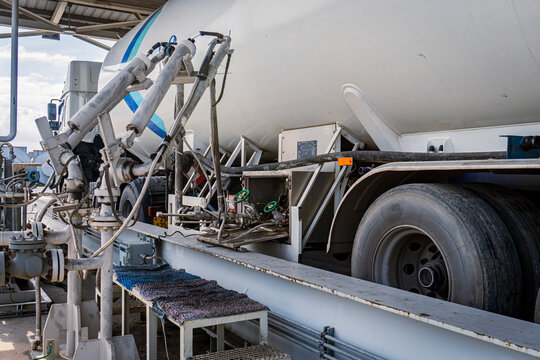 LNG or liquefied gas tank truck connected to a filling station, showing complex piping, valves, and hose connections along the side of the tanker at an industrial gas loading facility.