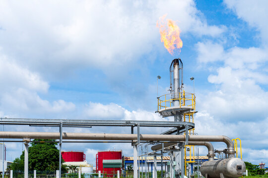 Large gas flare burning dramatically from the top of a flare stack against a blue sky, with foreground pipelines, red storage tanks, and yellow railings at an industrial plant facility.
