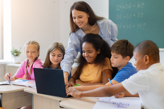 Female teacher assisting group of students using laptop at classroom desk, pointing at the screen during an interactive lesson, promoting technology, teamwork, and interactive learning