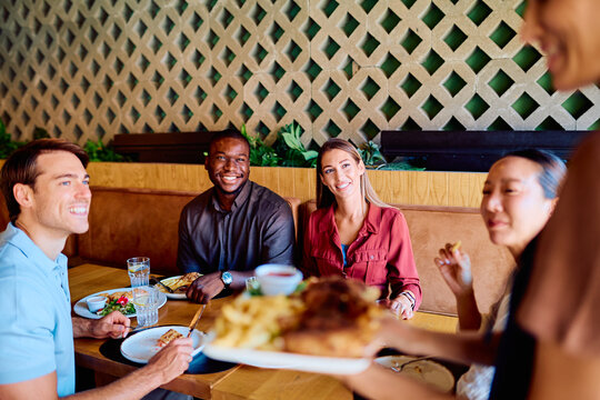 Diverse group of smiling friends enjoying a social gathering and lunch at a modern restaurant, being served by a waiter