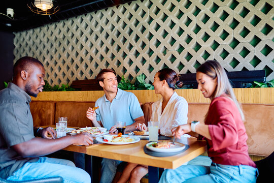 Diverse group of young adult friends smiling, eating, and socializing at a modern restaurant table, enjoying a shared meal