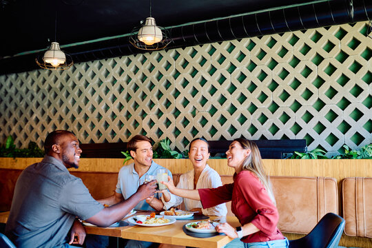 Diverse group of friends dining in a modern restaurant, making a cheerful toast with refreshing drinks, laughing and sharing happy moments