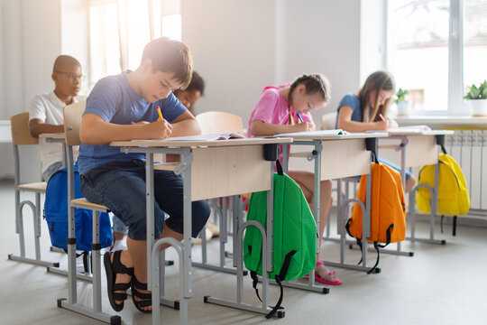 Diverse schoolchildren completing written assignments at individual desks in bright classroom, with colorful backpacks arranged beside their workstations