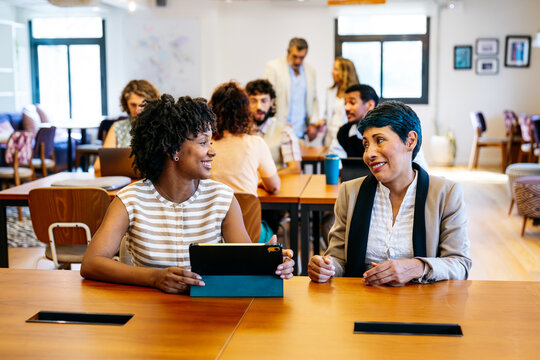 Two businesswomen discussing work on a tablet, surrounded by other professionals collaborating in a vibrant, shared workspace