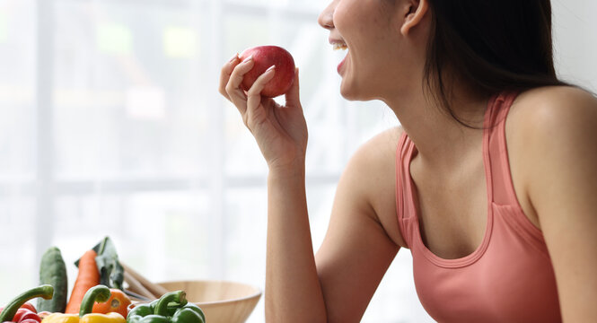Asian woman wearing a sports top, smiling widely and looking joyful while getting ready to take a bite from a fresh red apple.