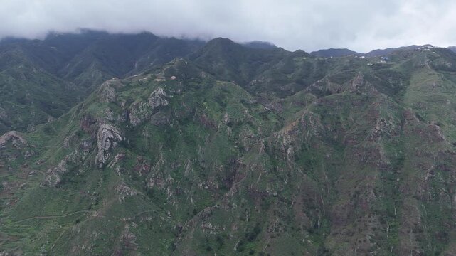 Aerial drone shot with a push in movement reveals the mountainous landscape near Taborno hamlet, nestled amidst the rugged, green mountains on Tenerife (Canary Islands), under a cloudy sky.