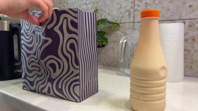Person placing a grocery paper bag on the kitchen counter and putting a bottle of fresh dairy product inside, showcasing the daily routine of shopping and home life