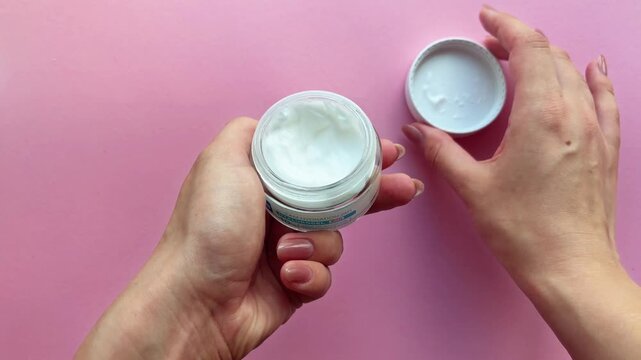 Woman with manicured nails opening a jar and applying moisturizing hand cream for skincare routine on a pink background, showcasing beauty product and self care