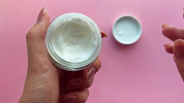 Woman's hands with a manicure holding an open glass jar of white cosmetic cream, scooping the product with a finger to test its texture over a plain pink background