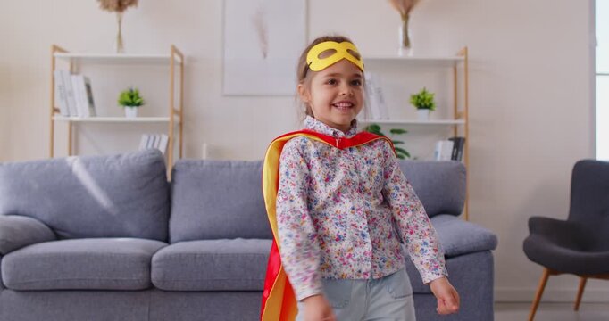 Cute preschooler 5s girl dressed like superhero in yellow fancy dress, cloak and mask dancing alone indoors, enjoy weekend games in living room. Carefree childhood, imagination, activity at home