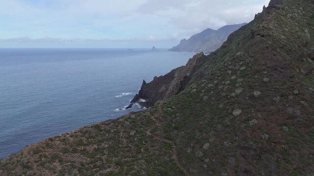 An aerial drone shot pans left along the dramatic, rugged coastline of Tenerife, Canary Islands revealing Roque de las Animas. Steep cliffs covered in sparse vegetation meet the blue Atlantic Ocean.