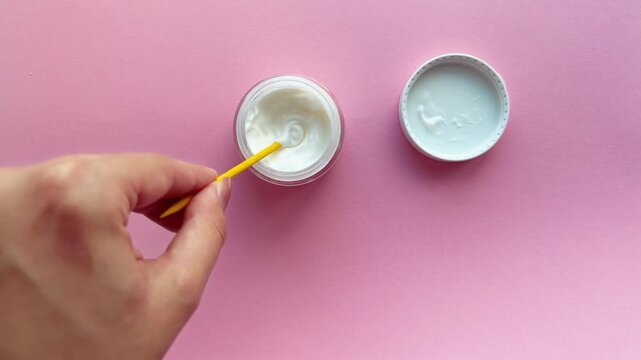 Woman's hand stirring and scooping white cosmetic skincare cream from a jar with a yellow spatula on a pink background, showcasing beauty product texture and consistency