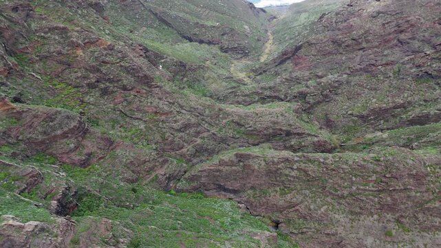 The drone booms down and pushes in, revealing a rugged mountain valley on Tenerife. Lush green vegetation and small huts dot the rocky cliffs, leading to the waves of Playa de Tamadite.