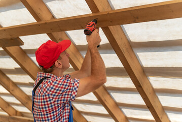 Male worker wearing a red cap and plaid shirt carefully drilling a screw into a wooden rafter, constructing the roof frame of a new house or renovation project © somemeans