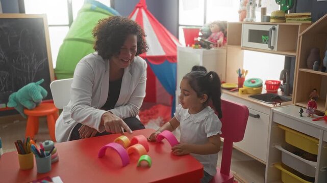 Woman points finger to colorful stacking blocks on red table in daycare building; nurturing learning play.