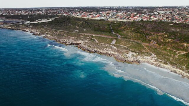 Turquoise ocean waves crash against the rocky shoreline near the green landscape of Burns Beach in Perth, Australia on a bright summer afternoon.