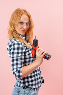 Confident young woman with red hair wearing a plaid shirt and safety glasses, holding a cordless electric screwdriver, ready for home improvement work