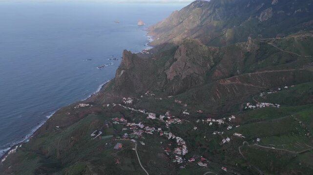 Aerial pan left drone shot captures the rugged coast and lush mountains on Tenerife, home to the village Taganana and Roque de las &Aacute;nimas. A stunning sunset illuminates the dramatic landscape.