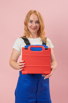 Woman in blue overalls smiling and holding a red tool case with both hands, presenting equipment for home repair, construction, or DIY projects