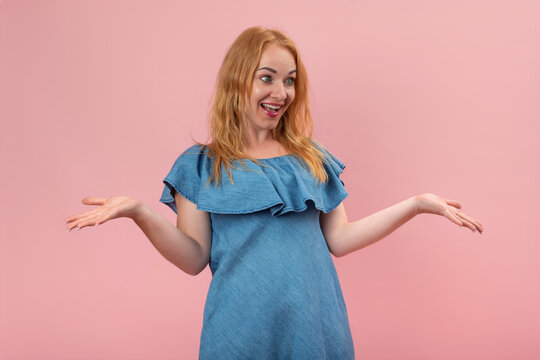 Young woman with red hair looking to the side, portraying surprise and amazement while shrugging and presenting something with open palms on a pink background, expressing an emotional reaction