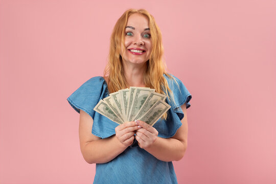 Happy woman with wide eyes and a big smile holding a fan of one hundred dollar bills, expressing excitement and abundance on a pink background
