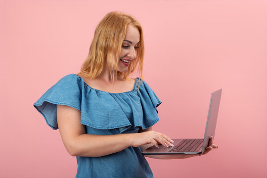 Blonde young woman happily using a laptop, typing on the keyboard with a joyful expression, engaging with online content or communicating casually against a pink background